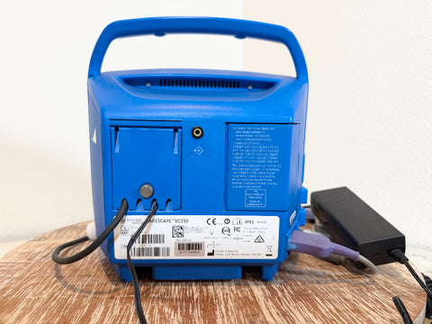 The back of a blue VitalSigns VC150 portable blood oxygen monitor, shown on a wooden surface with its power adapter plugged in against a plain white wall.
