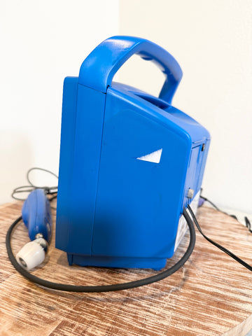 A bright blue VitalSigns VC150 portable blood oxygen monitor, shown from a side angle on a wooden surface with its black power cord and hand grip visible.