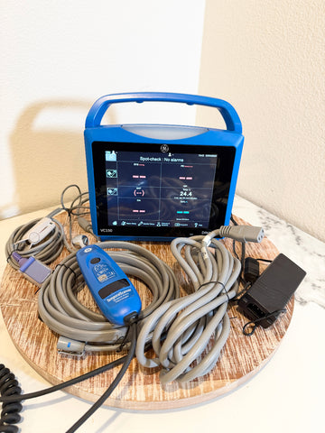 A VitalSigns VC150 portable blood oxygen monitor with a blue handle and screen, displayed on a round wooden tray with attached gray cables, a blue handheld controller, and a black power adapter against a beige wall and white marble surface.