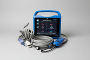 A handheld VitalSigns VC150 portable blood oxygen monitor with a blue handle and screen, shown with coiled gray cables and a matching blue handheld unit on a round wooden tray against a beige wall and marble surface.