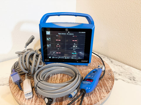 A handheld VitalSigns VC150 portable blood oxygen monitor with a blue handle and screen, shown with coiled gray cables and a matching blue handheld unit on a round wooden tray against a beige wall and marble surface.