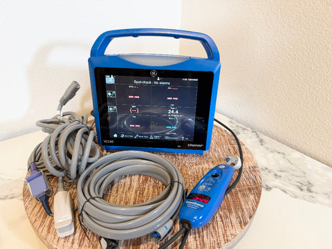 A GE VitalSigns VC150 portable blood oxygen monitor with a blue handle and screen, shown with gray cables and a pulse oximeter on a round wooden tray against a beige wall.