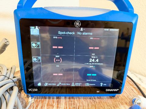 A blue GE Dinamap VC150 portable medical monitor with a touchscreen display, resting on a wooden surface with cables visible.