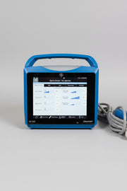 A blue VitalSigns VC150 portable blood oxygen monitor with a handle and screen, sitting on a wooden table with a coiled cable and a blue handheld attachment nearby.