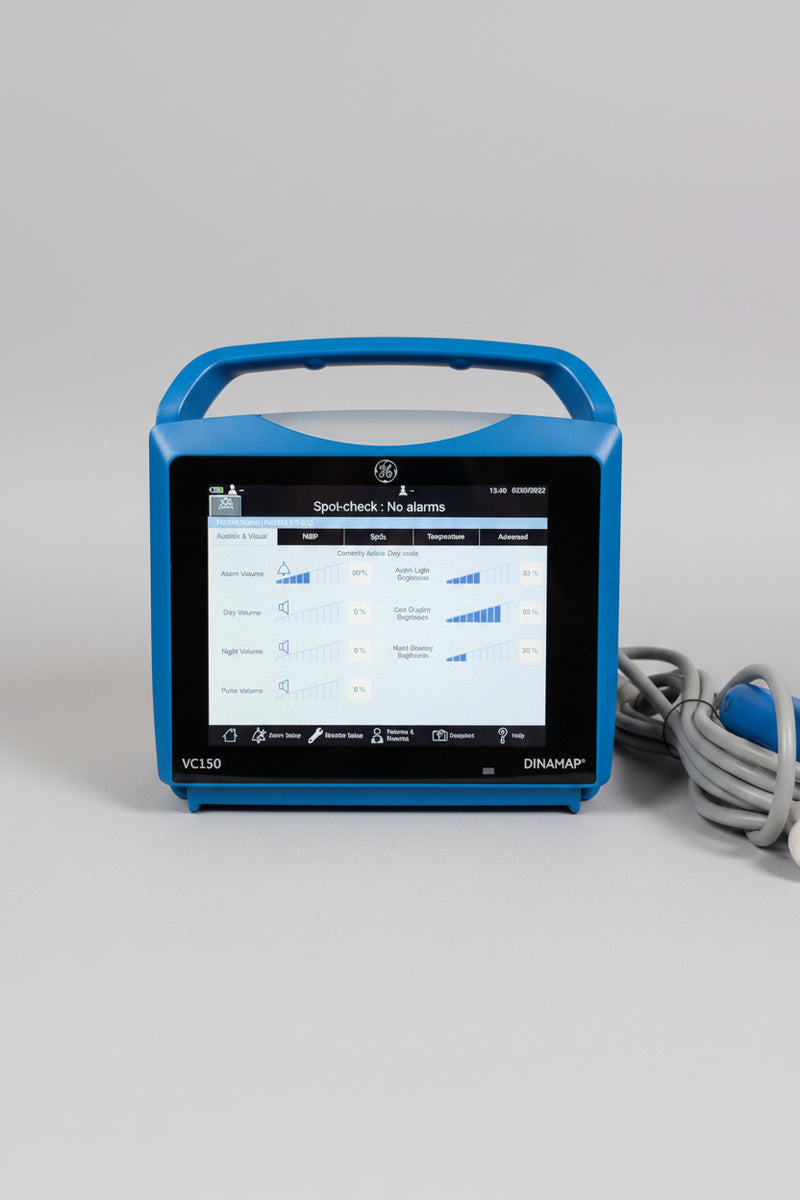A blue VitalSigns VC150 portable blood oxygen monitor with a handle and screen, sitting on a wooden table with a coiled cable and a blue handheld attachment nearby.