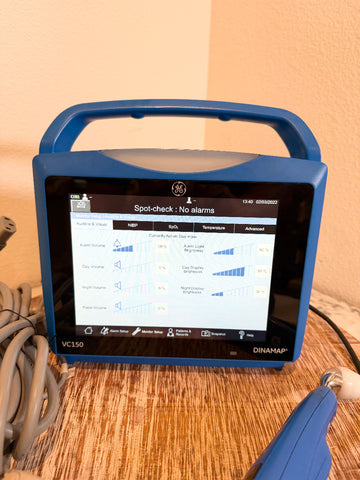 A blue VitalSigns VC150 portable blood oxygen monitor with a handle and screen, sitting on a wooden table with a coiled cable and a blue handheld attachment nearby.