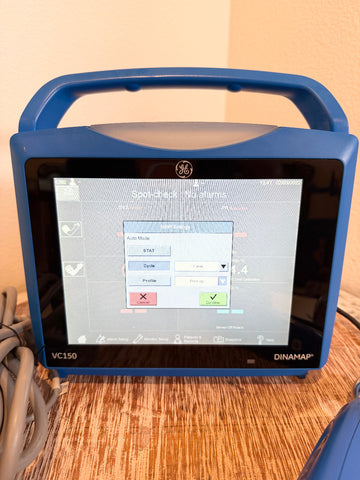 A blue VitalSigns VC150 portable blood oxygen monitor with a handle and touchscreen display, sitting on a wooden surface.