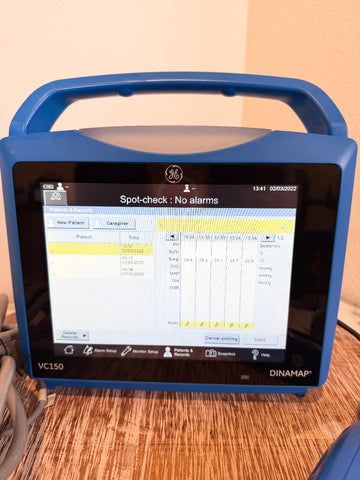 A GE Dinamap VC150 portable medical monitor with a blue casing and handle, showing a patient monitoring screen on a wooden tabletop.