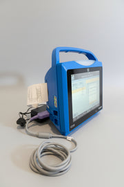 A blue VitalSigns VC150 portable blood oxygen monitor with a screen display, placed on a woven mat with cables and a paper printout visible.