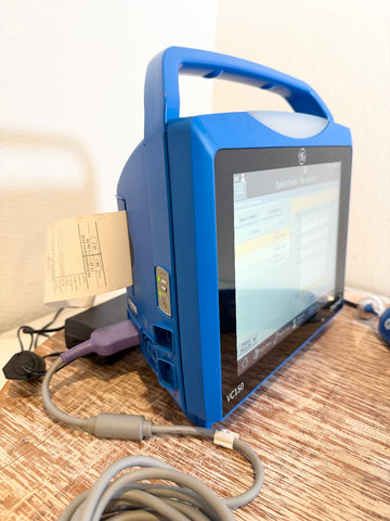 A blue VitalSigns VC150 portable blood oxygen monitor with a screen display, placed on a woven mat with cables and a paper printout visible.