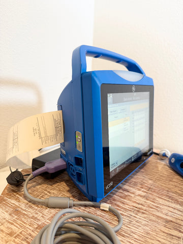 A blue VitalSigns VC150 portable blood oxygen monitor with an integrated handle and screen, shown on a wooden tabletop with a coiled cable and paper printout attached. 