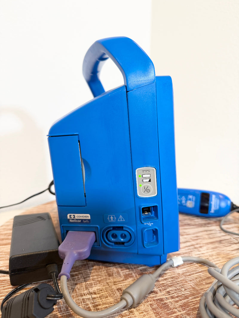 A blue VitalSigns VC150 portable blood oxygen monitor shown from the back, with its power cord plugged in, resting on a wooden table.