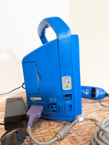 A blue VitalSigns VC150 portable blood oxygen monitor shown from the back, with its power cord plugged in, resting on a wooden table.