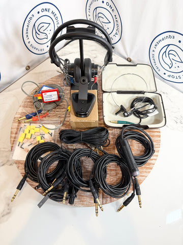 A set of audio equipment laid out on a round wooden tray, including a pair of over-ear headphones on a stand, multiple coiled audio cables with 3.5mm and jack connectors, a small control module, yellow earplugs, and a white storage case.