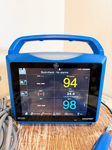 A blue GE Dinaram VC150 pulse oximeter sitting on a wooden table against a beige wall, showing a spot-check screen with readings of 94 and 98.