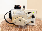 A KMI flow meter pump with a cream-colored metal body, black knobs, and a black power cord, resting on a round wooden surface against a plain beige wall.