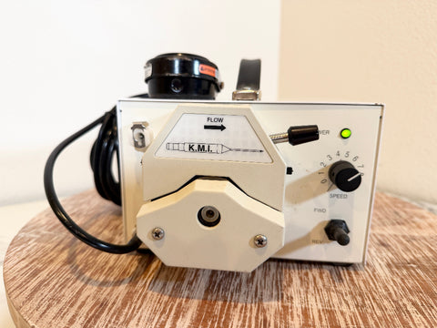 A KMI flow meter pump with a cream-colored metal body, black knobs, and a black power cord, resting on a round wooden surface against a plain beige wall.