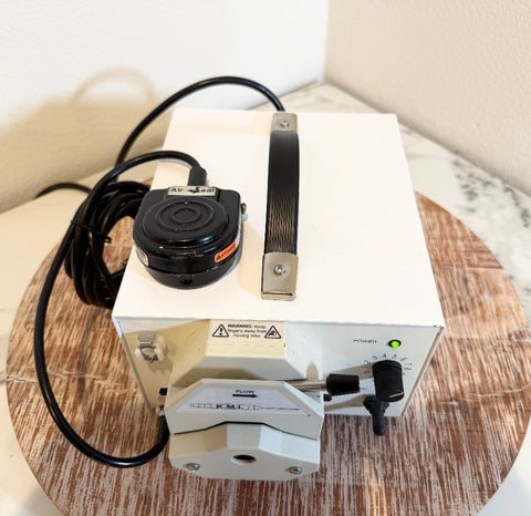 A white and black KMI flow meter pump with a carrying handle and foot pedal, resting on a round wooden tray.