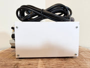 A white rectangular metal box with four corner screws, topped with a coiled black power cord, sitting on a wooden surface against a plain beige wall.