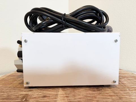 A white rectangular metal box with four corner screws, topped with a coiled black power cord, sitting on a wooden surface against a plain beige wall.
