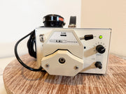 A KMI flow meter with a cream-colored metal body, black dials, and a black cable, resting on a round wooden surface against a plain beige wall.