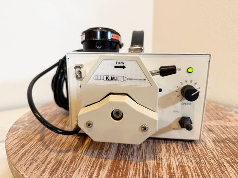 A KMI flow meter with a cream-colored metal body, black dials, and a black cable, resting on a round wooden surface against a plain beige wall.