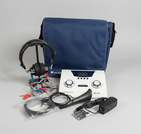 A Maico MA25 audiometry testing kit laid out on a round wooden tray, including a white and blue electronic device, black headphones, a black microphone, cables, an adapter, and a blue carrying case against a white wall background.