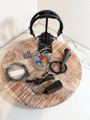 Maico MA25 accessories displayed on a round wooden board, including a black over-ear headset on a stand, a black joystick, cables, connectors, a power adapter, and a small battery pack against a white marble-patterned surface.