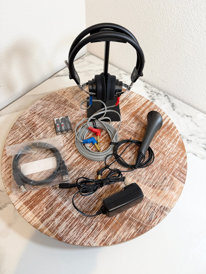 Maico MA25 accessories displayed on a round wooden board, including a black over-ear headset on a stand, a black joystick, cables, connectors, a power adapter, and a small battery pack against a white marble-patterned surface.