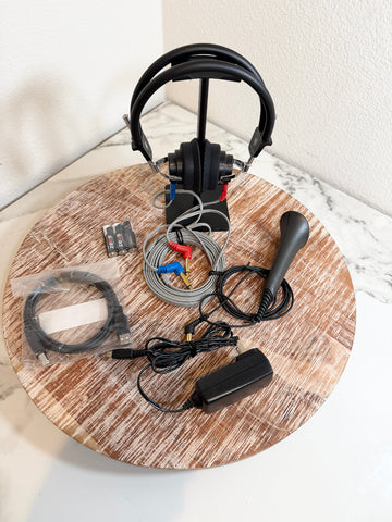 Maico MA25 accessories displayed on a round wooden board, including a black over-ear headset on a stand, a black joystick, cables, connectors, a power adapter, and a small battery pack against a white marble-patterned surface.