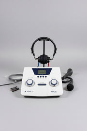 A MAICO MA 25 hearing testing device with a white faceplate, blue accents, and an LCD screen, displayed on a round wooden tray with its accessories including headphones, a microphone, and batteries.