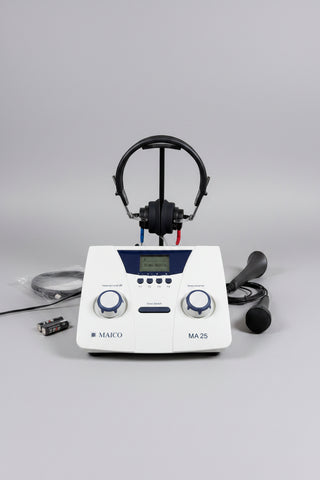 A MAICO MA 25 hearing testing device with a white faceplate, blue accents, and an LCD screen, displayed on a round wooden tray with its accessories including headphones, a microphone, and batteries.