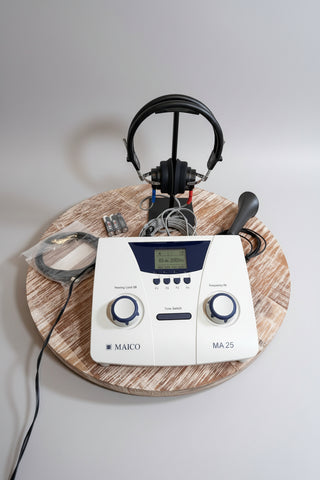 A MAICO MA25 hearing testing device with a white front panel, blue accents, and a digital display, shown with attached headphones, ear molds, and a probe on a round wooden tray against a white wall.