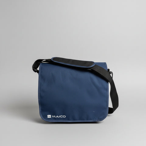 A navy blue Maico messenger bag with a black shoulder strap, resting on a round wooden stool against a plain off-white wall.