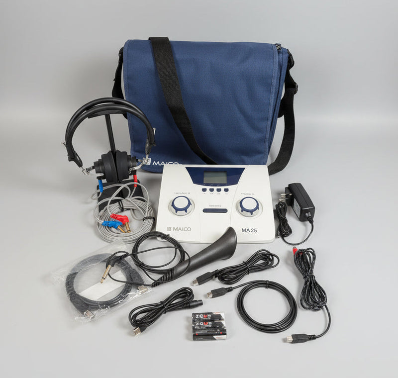 A Maico MA25 device kit displayed on a round wooden tray, including the white and blue control unit, black headphones, speaker, cables, battery, charger, and a blue carrying bag against a white wall.