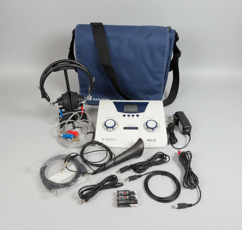 A Maico MA25 device kit displayed on a round wooden tray, including the white and blue control unit, black headphones, speaker, cables, battery, charger, and a blue carrying bag against a white wall.