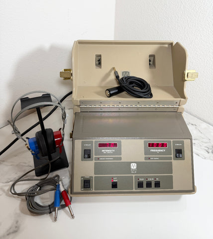 A Maico MA39 Portable Diagnostic Audiometer device with its open hood, connected to headphones, ear tubes, and cables on a white marble surface against a white wall.