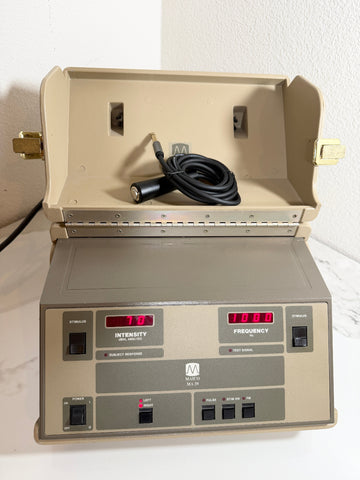 A Maico MA39 Portable Diagnostic Audiometer device with an open beige carrying case, a coiled black cable inside, and a front control panel with red digital displays and switches. The device is photographed on a white surface against a white wall.