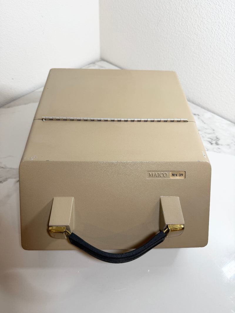 A Maico MA39 Portable Diagnostic Audiometer beige case with a black handle and gold accents, sitting on a white marble-patterned surface against a white wall.
