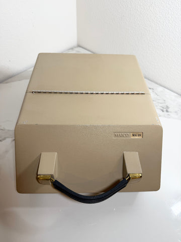 A Maico MA39 Portable Diagnostic Audiometer beige case with a black handle and gold accents, sitting on a white marble-patterned surface against a white wall.