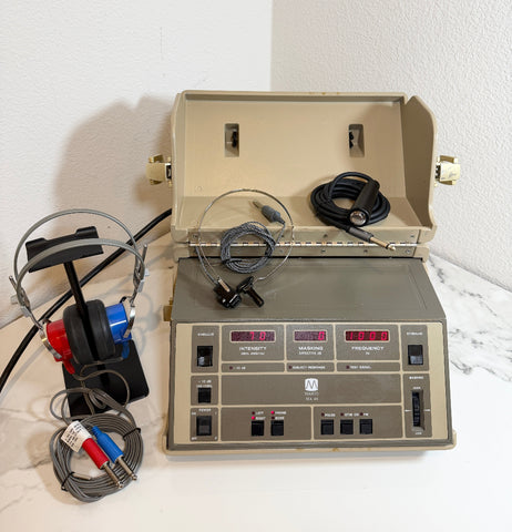 A Maico MA40 Portable Diagnostic Audiometer with its open beige case, attached gray and black cables, and a pair of red-and-blue headphones on a white marble surface against a white wall.