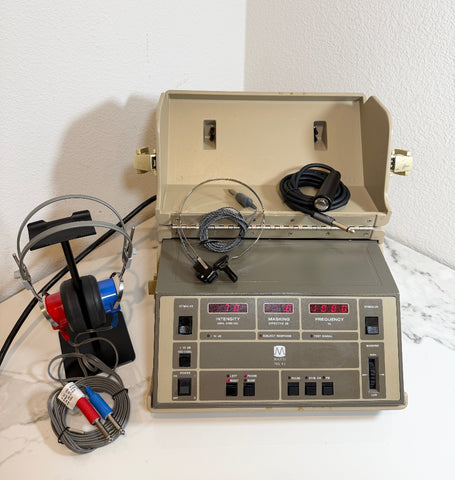 A Maico MA40 Portable Diagnostic Audiometer with an open beige case, control panel with digital displays and switches, attached cables, and a pair of over-ear headphones on a stand, photographed against a white wall and light surface.