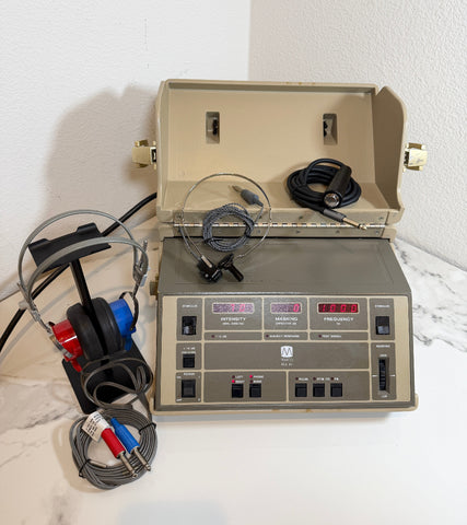 A Maico MA40 Portable Diagnostic Audiometer with an open beige cover, a front control panel with switches and digital displays, attached gray cables, and a pair of over-ear headphones on a stand, photographed on a white surface against a white wall.