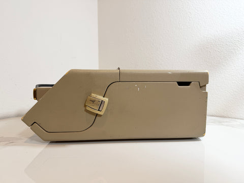 A beige Maico MA40 Portable Diagnostic Audiometer with a gold-toned latch, shown from a side profile on a white surface against a white wall.