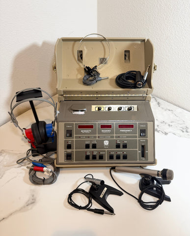 A Maico MA41 Portable Diagnostic Audiometer device with its open carrying case, shown with attached and nearby headphones, ear pieces, and a handheld microphone on a white marble surface against a white wall.