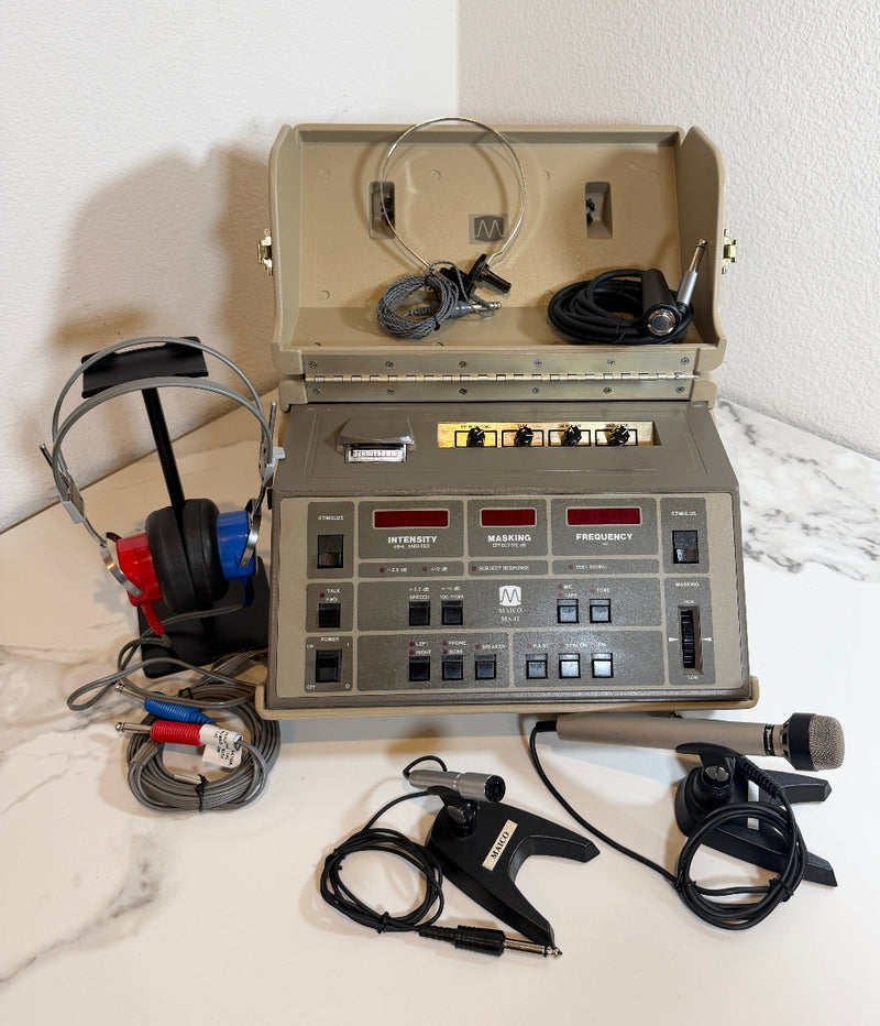 An open beige pMaico MA41 Portable Diagnostic Audiometer device with a front control panel, shown on a white marble-like surface against a white wall. Attached accessories include over-ear headphones, a microphone on a stand, and cables, with the main unit open to reveal internal cords and components.