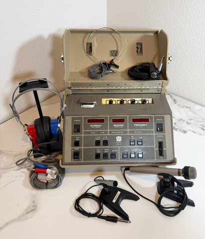 An open beige pMaico MA41 Portable Diagnostic Audiometer device with a front control panel, shown on a white marble-like surface against a white wall. Attached accessories include over-ear headphones, a microphone on a stand, and cables, with the main unit open to reveal internal cords and components.