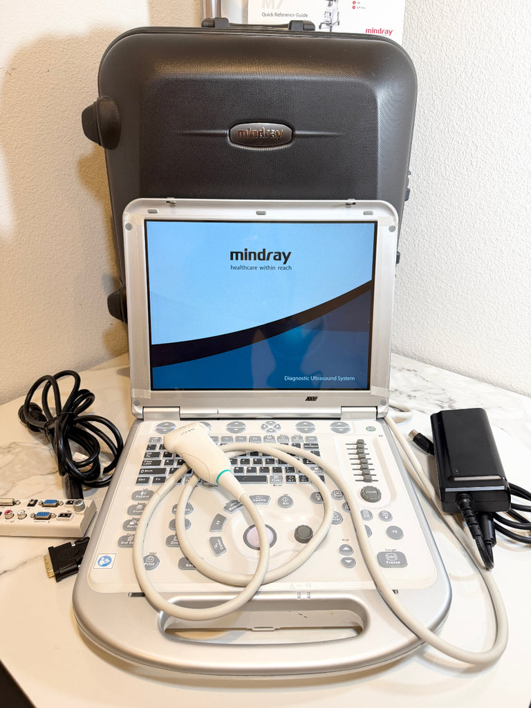 A Mindray portable ultrasound machine with its control console, probe cable, power adapter, remote, and carrying case on a white surface against a white wall.