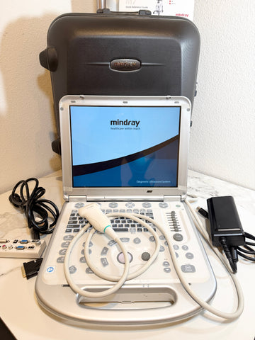 A Mindray portable ultrasound machine with its control console, probe cable, power adapter, remote, and carrying case on a white surface against a white wall.