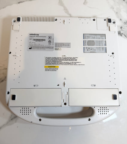 The underside of a white Mindray handheld ultrasound device, showing the brand label, ventilation grilles, and handle cutout, photographed on a white marble surface.